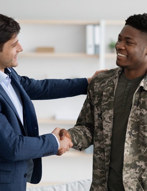 Cheerful guy in blue suit shaking happy young black military man hand, psychologist and soldier celebrating successful therapy, clinic interior. Military rehabilitation service concept